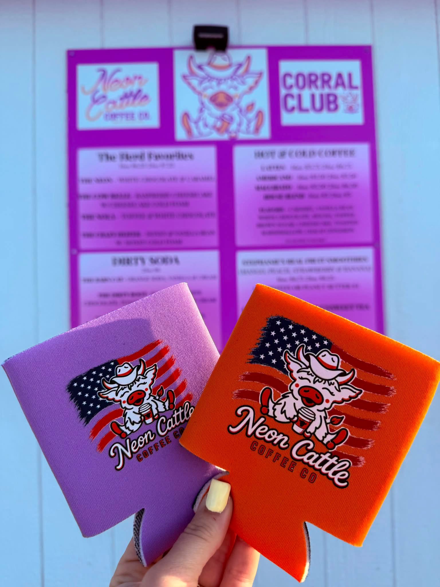 Purple and orange Neon Cattle koozies with a flag-and-cow design, held up in front of the menu board