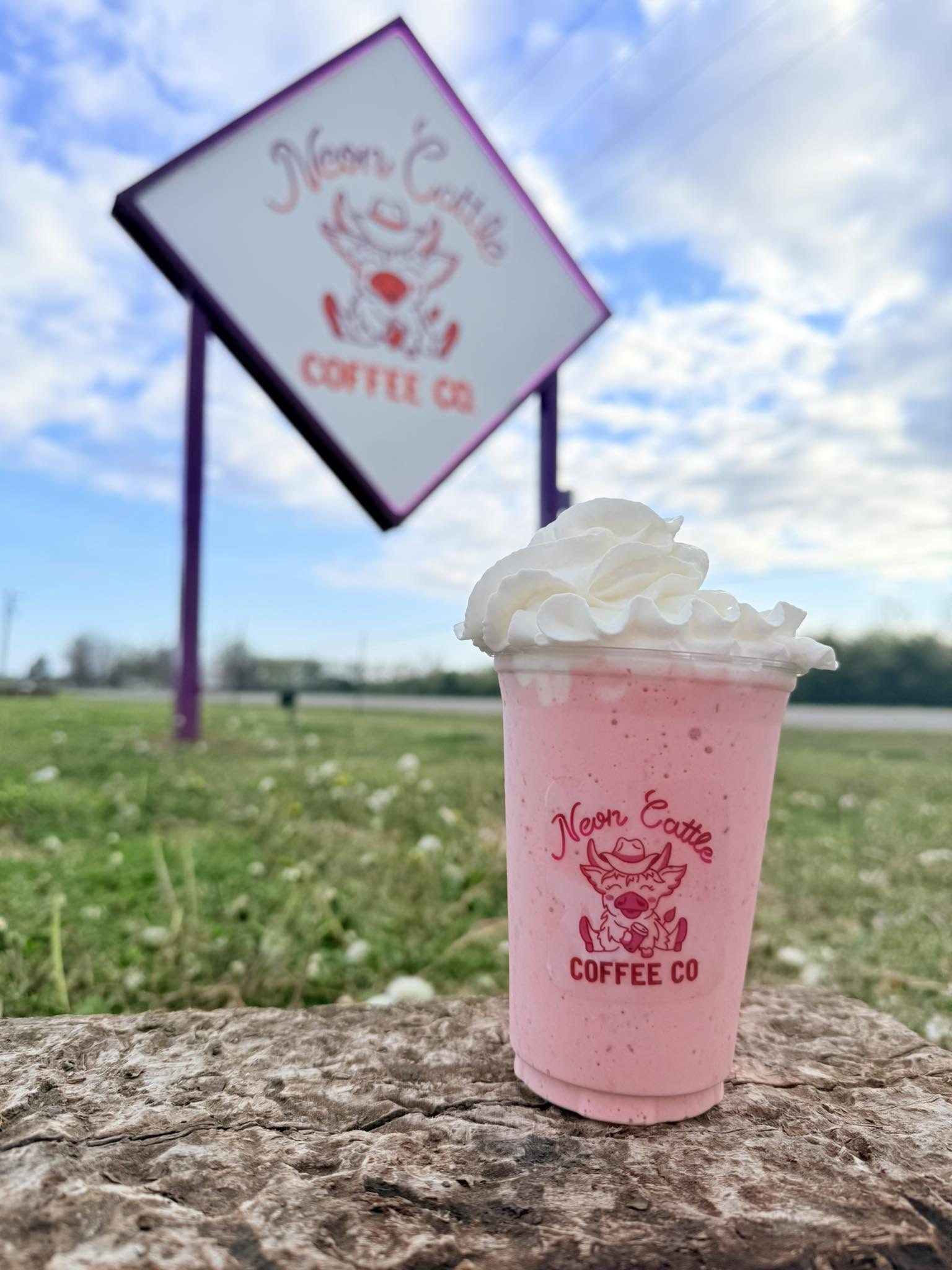 Pink blended drink topped with whipped cream in a Neon Cattle cup, with the roadside sign behind it